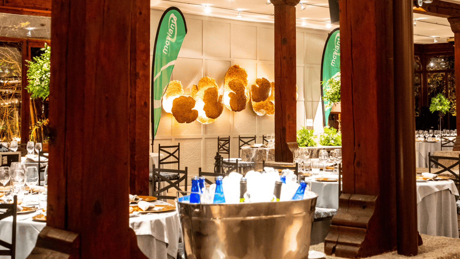 Salón decorado para un evento con mesas elegantes, botellas de agua en hielo y banderas de Moriana.
