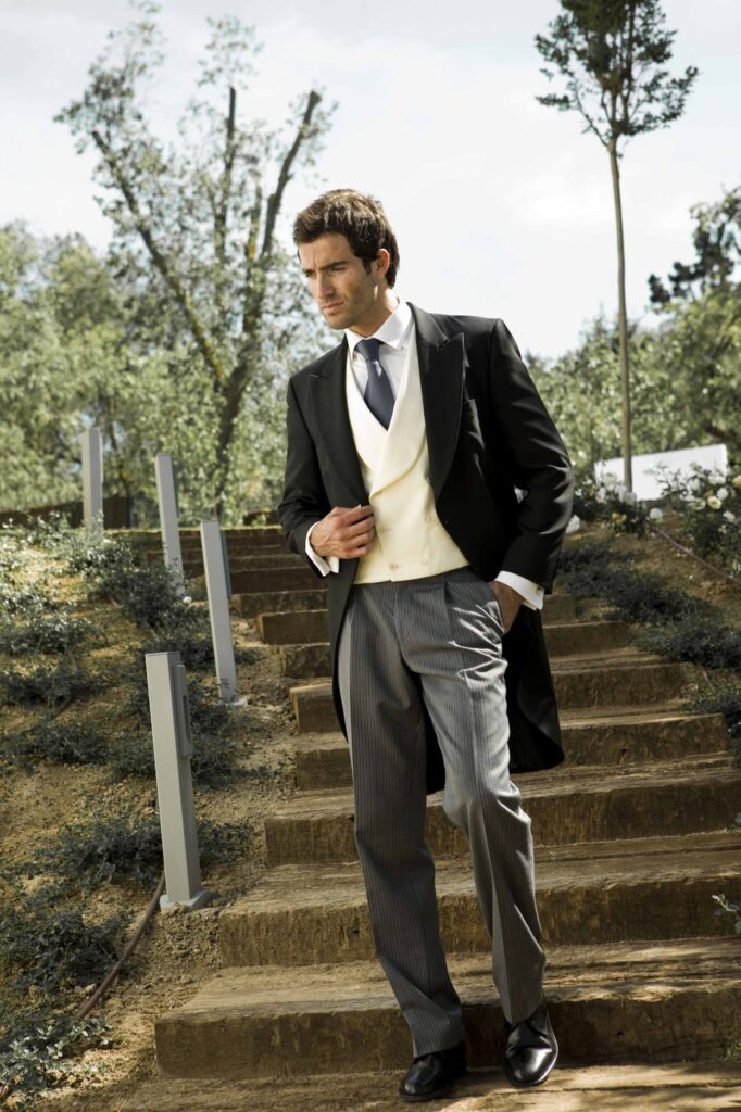 Hombre elegante con chaqué negro, chaleco crema y corbata gris, caminando por escaleras al aire libre.