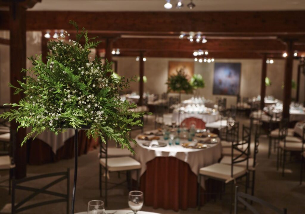 Salón de eventos decorado con mesas redondas y centros de mesa con flores y hojas verdes.