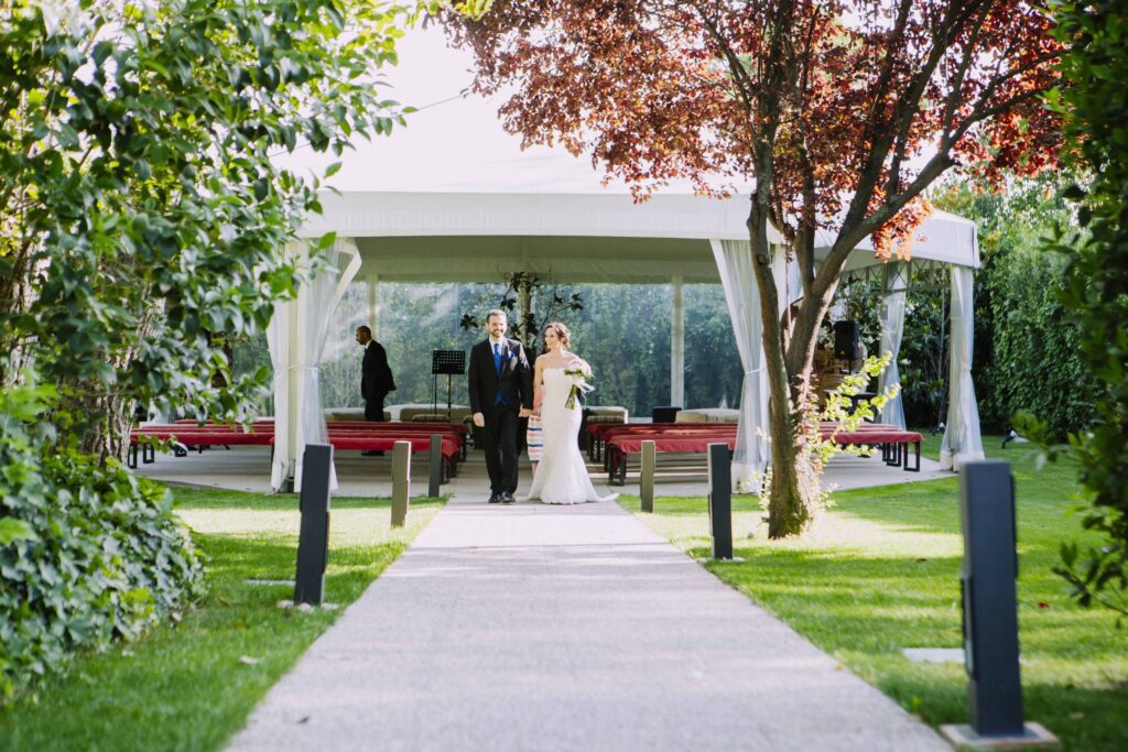 Pareja de recién casados caminando por un sendero de jardín bajo una carpa blanca en La Quinta de Illescas.