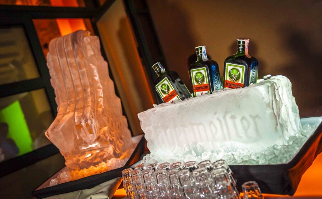 Estación de Jägermeister en La Quinta de Illescas con una escultura de hielo personalizada que sostiene botellas de licor Jägermeister, rodeada de copas de cristal listas para servir en un evento elegante y moderno.
