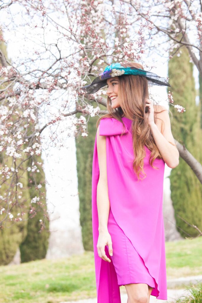 Invitada con vestido fucsia y pamela posando junto a árboles en flor en la Quinta de Illescas