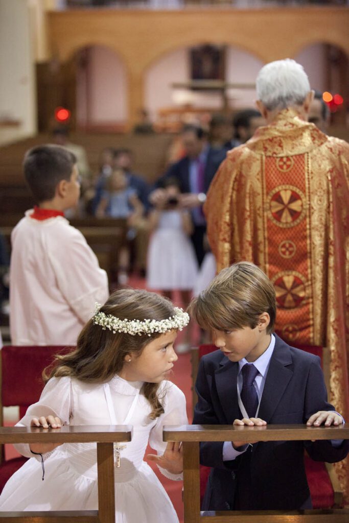 Niños vestidos de blanco y traje oscuro, en ceremonia religiosa, hablando en el altar.