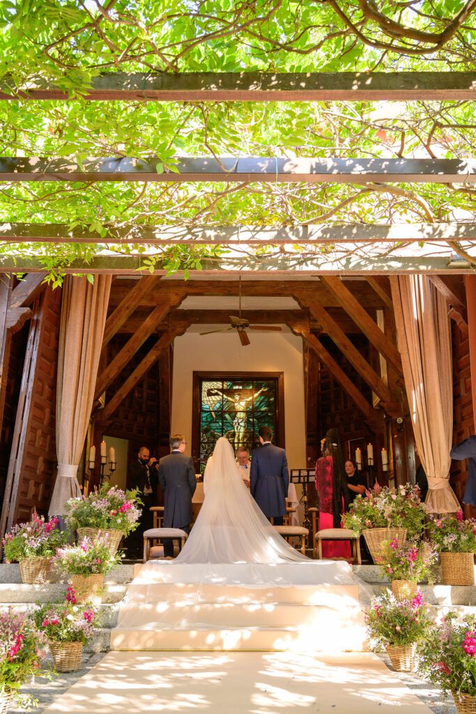 Ceremonia religiosa bajo una pérgola de madera con vegetación y vidriera al fondo en La Quinta de Illescas