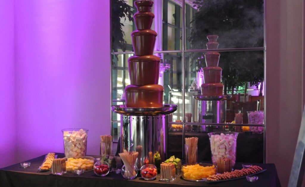 Estación de fuente de chocolate en un evento de La Quinta de Illescas, acompañada de recipientes con frutas frescas, malvaviscos y barquillos, iluminada por luces púrpuras. La fuente de varios niveles crea una atmósfera lujosa y deliciosa para los invitados.