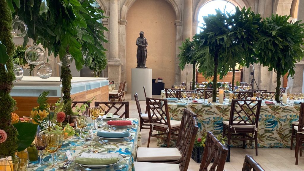 Mesa imperial con vajilla, copas doradas y mantelería estampada, rodeada de centros florales tropicales en el claustro del Museo del Prado durante un evento de La Quinta de Illescas