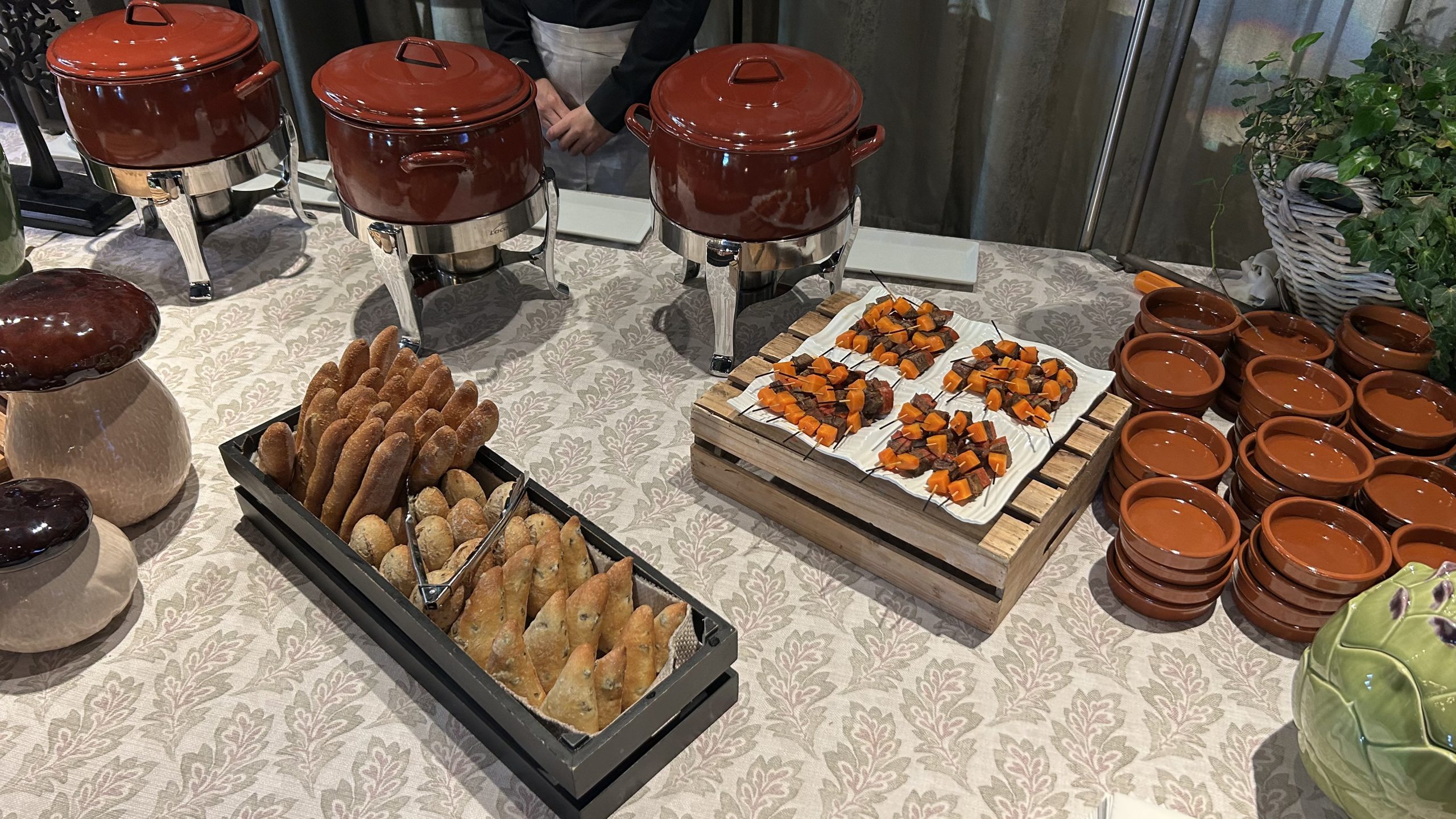Bufet de guisos tradicionales para bodas con tres ollas de puchero rojas, bandejas de pan y brochetas.